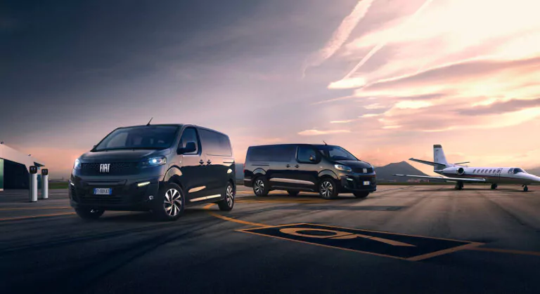 Zwei schwarze Fiat-Kleintransporter stehen bei Sonnenuntergang auf einer Landebahn eines Flughafens. Im Hintergrund ist ein weißer Privatjet zu sehen und die Berge sind unter einem dramatischen Himmel zu erkennen.