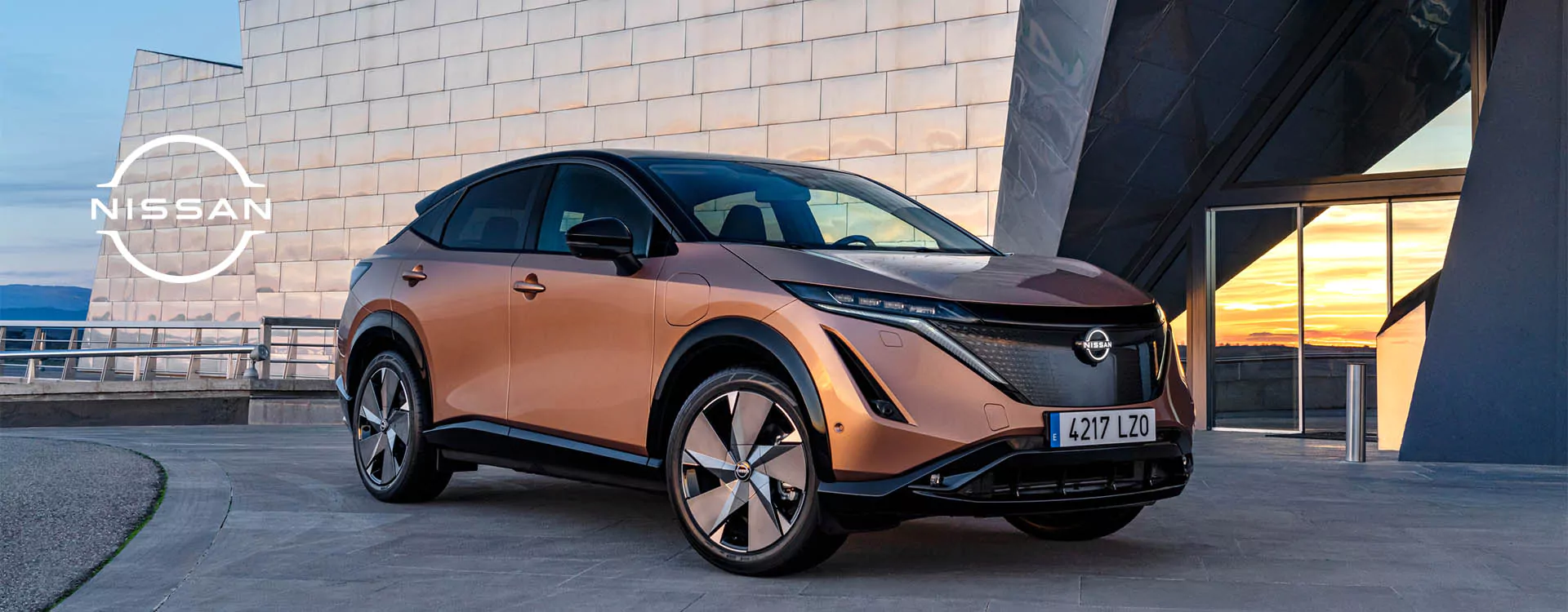 Ein bronzener Nissan Ariya Elektro-SUV ist bei Sonnenuntergang vor einem modernen Glasgebäude geparkt, wobei das Nissan-Logo auf der linken Seite des Bildes zu sehen ist.