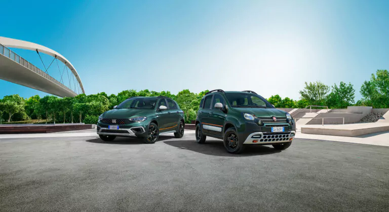 Zwei grüne Fiat-Autos sind auf einem gepflasterten Platz im Freien geparkt, in der Nähe einer modernen weißen Brücke und üppig grünen Bäumen unter einem klaren blauen Himmel.