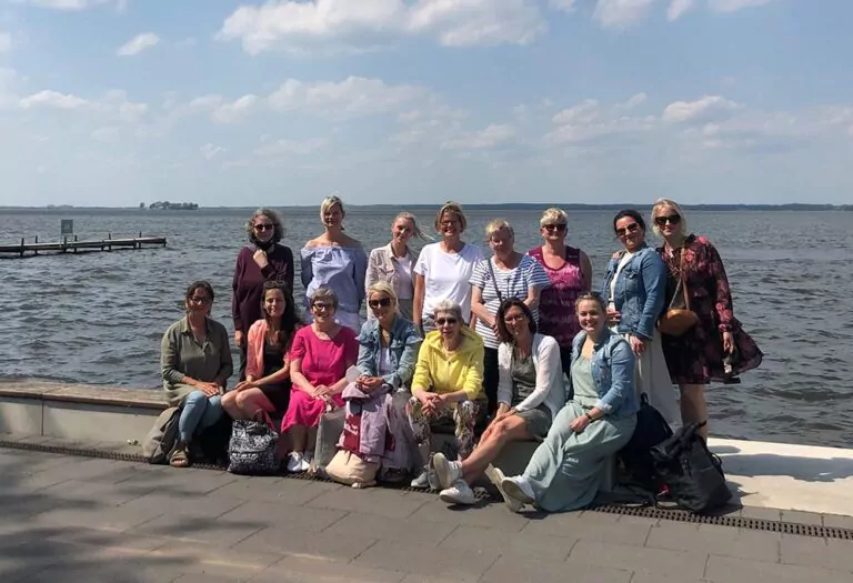 Eine Gruppe von 14 Frauen posiert gemeinsam auf einem gepflasterten Gehweg an einem großen Gewässer unter einem teilweise bewölkten Himmel. Einige sitzen, andere stehen, und sie alle scheinen den sonnigen Tag zu genießen.