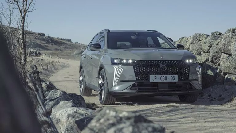 Ein silberner Geländewagen fährt auf einer unbefestigten Straße, umgeben von Felsen und trockener Landschaft, unter einem klaren Himmel. Das Fahrzeug hat moderne Scheinwerfer und einen markanten Kühlergrill mit einem sichtbaren Nummernschild.