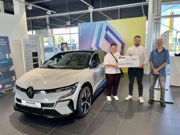 Drei Personen stehen neben einem weißen Renault Megane in einem Ausstellungsraum. Der Mann in der Mitte hält einen großen Scheck über 2.000 Euro in der Hand. Im Hintergrund sind bunte Schilder und große Fenster zu sehen.