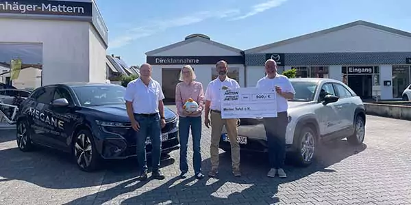 Vier Personen stehen vor zwei Autos vor einem Gebäude. Eine Person hält einen Fußball, eine andere einen großen Spendenscheck über 500 Euro. Es ist sonnig und die Gruppe lächelt in die Kamera.