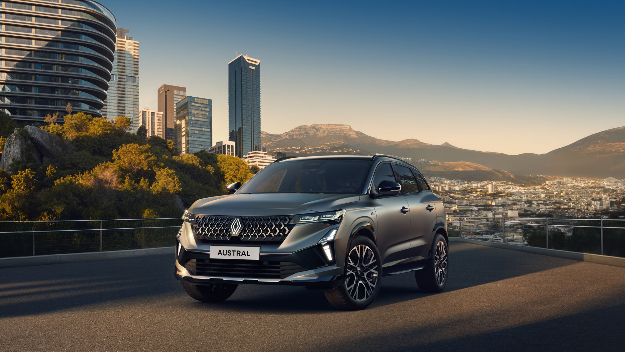 Ein silberner Renault Austral SUV, der über Renault Austral Leasing erhältlich ist, parkt auf einer Straße mit Blick auf eine moderne Stadt mit hohen Gebäuden und fernen Bergen unter einem klaren Himmel.