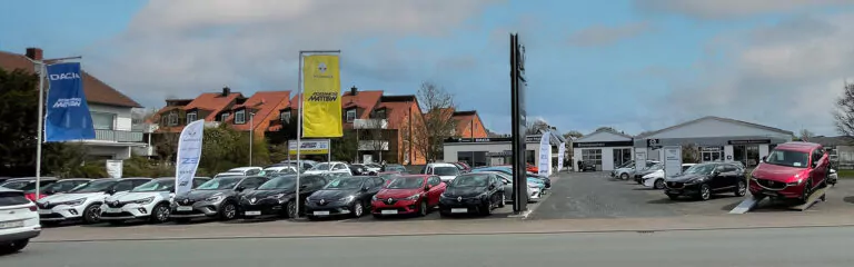 Ein Autohaus mit Reihen von geparkten Fahrzeugen, Autohausflaggen und Gebäuden im Hintergrund. Ein rotes Auto steht auf einer Plattform in der Nähe des Vordereingangs, und der Himmel ist teilweise bewölkt.