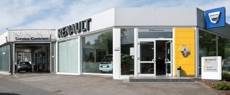 Außenansicht eines Renault-Händlers und -Servicezentrums mit großen Glasfenstern, sichtbaren Autos im Inneren, einem Willkommen-Schild am Eingang und einem Dacia-Logo am Gebäude.