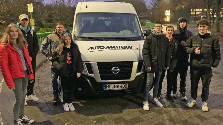 Eine Gruppe von neun Teenagern in Jacken und Freizeitkleidung, einige mit BTW-Basketball-Ausrüstung, steht um einen weißen Nissan-Van mit Auto-Mattern auf der Motorhaube, der abends auf einer Kopfsteinpflasterstraße geparkt ist.