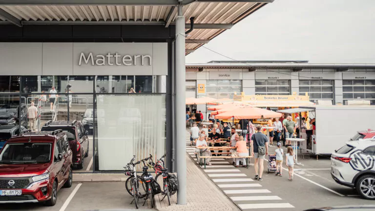 Auf einem belebten Markt unter freiem Himmel tummeln sich Menschen, die das Wochenende genießen und sich um Essensstände und Picknicktische scharen. In der Nähe reihen sich geparkte Autos und Fahrräder vor einem modernen Gebäude mit der Aufschrift Mattern auf. Die Szene ist lebhaft und hell.