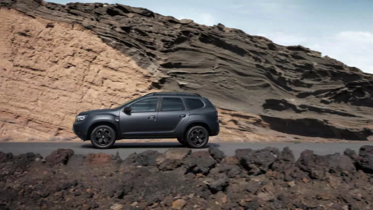 Ein dunkelgrauer Geländewagen parkt auf einer asphaltierten Straße mit zerklüfteten, geschichteten Felsformationen im Hintergrund unter einem teilweise bewölkten Himmel.