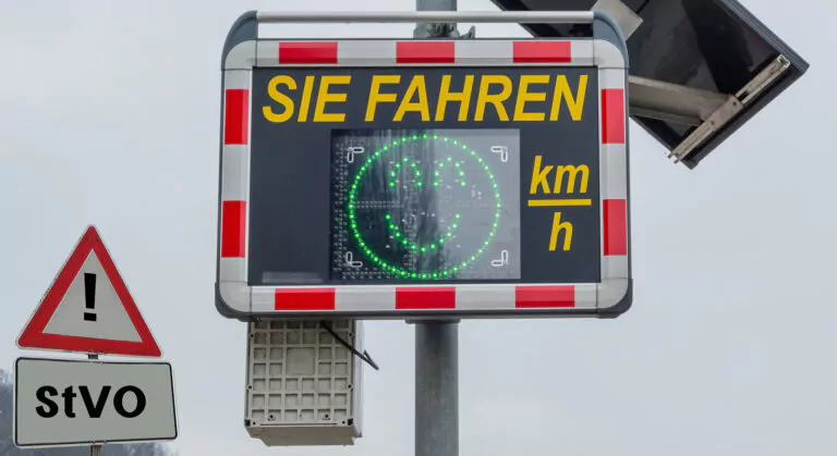Ein deutsches Geschwindigkeitsüberwachungsschild zeigt SIE FAHREN mit einem digitalen Smiley und km/h. Darunter sind ein dreieckiges Warnschild mit einem Ausrufezeichen und ein rechteckiges Schild mit der Aufschrift StVO zu sehen.