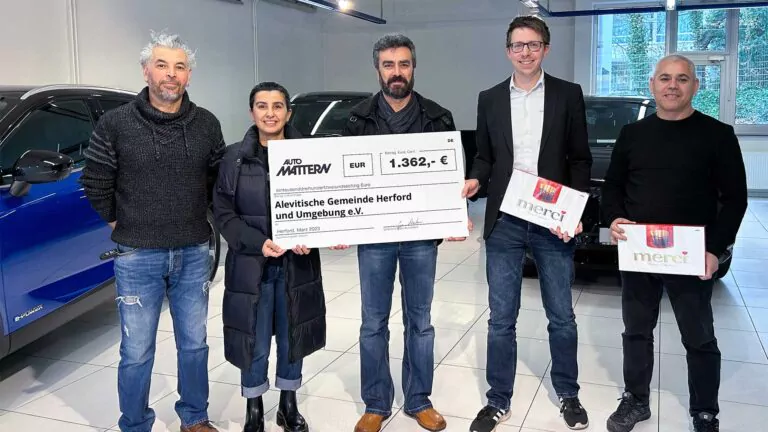 Fünf Personen stehen drinnen, zwei halten Merci-Schokoladenschachteln, die drei in der Mitte halten einen großen Spendenscheck über 1.362 Euro von Auto Matjev für die Spende Türkei Erdbeben Herford, umgeben von Autos.