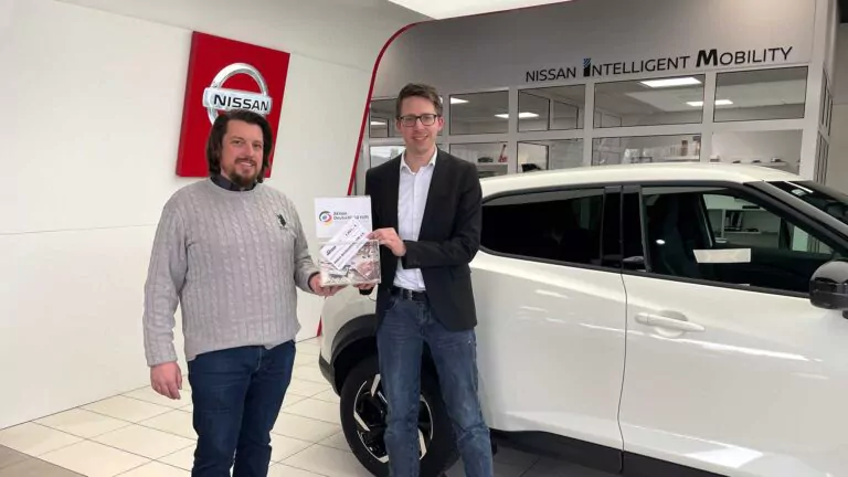 Zwei Männer stehen lächelnd neben einem weißen Nissan-Geländewagen in einem Autohaus, während der eine dem anderen eine Urkunde für Spenden Türkei Syrien überreicht. An der Wand hinter ihnen steht "NISSAN INTELLIGENT MOBILITY" und ein Nissan-Logo.