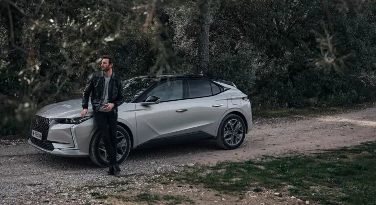 Ein Mann in einer schwarzen Jacke lehnt an einem silbernen Geländewagen, der auf einem von Bäumen und Grün umgebenen Feldweg geparkt ist.