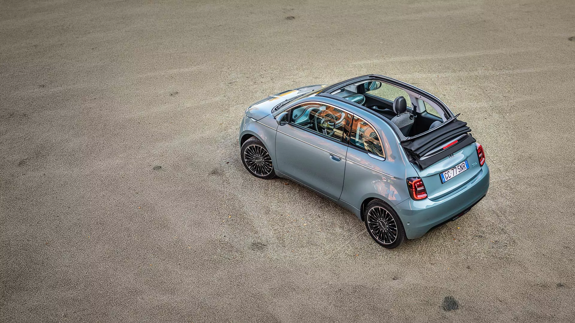 Ein hellblaues Fiat 500e Elektro Cabrio mit teilweise geöffnetem Verdeck ist auf einer sandigen Fläche geparkt, von oben und von hinten gesehen. Das Innere des Fahrzeugs und die schwarzen Leichtmetallräder sind sichtbar.