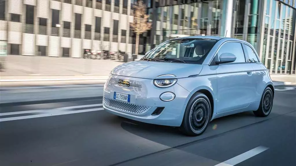 Ein hellblauer Fiat 500 Elektro fährt auf einer Stadtstraße mit modernen Glasgebäuden im Hintergrund, aufgenommen in Bewegung während des Tages.