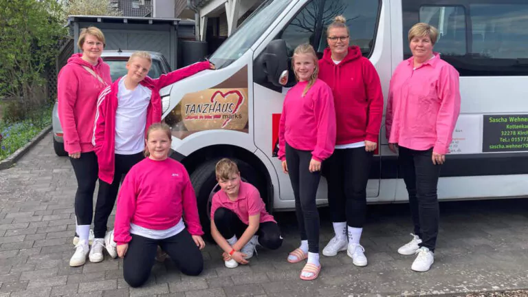 Sieben Personen, meist Frauen und Mädchen, in rosa und weißer Kleidung, stehen und knien neben einem weißen Lieferwagen mit dem Logo der Tanzschule Tanzhaus Marks LittleSteps und lächeln in die Kamera auf einem gepflasterten Außengelände.