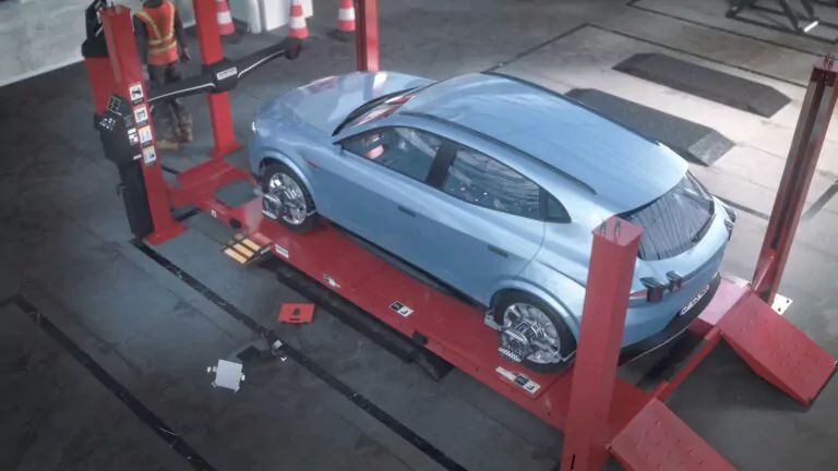 Ein blaues Auto steht auf einer roten Hebebühne in einer Autowerkstatt und wird einer Achsvermessung unterzogen. Eine Person mit einer orangefarbenen Sicherheitsweste steht in der Nähe, und im Hintergrund sind Verkehrskegel zu sehen.