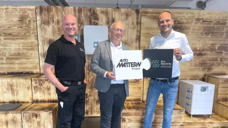 Drei lächelnde Männer stehen in einem Raum und halten zwei Firmenschilder in der Hand: auf dem einen steht Auto Mattern Gruppe, auf dem anderen Pro Electricity. Sie stehen vor einer Holzwand, die ihren gemeinsamen Fokus auf Erneuerbare Energien und Photovoltaik verdeutlicht.