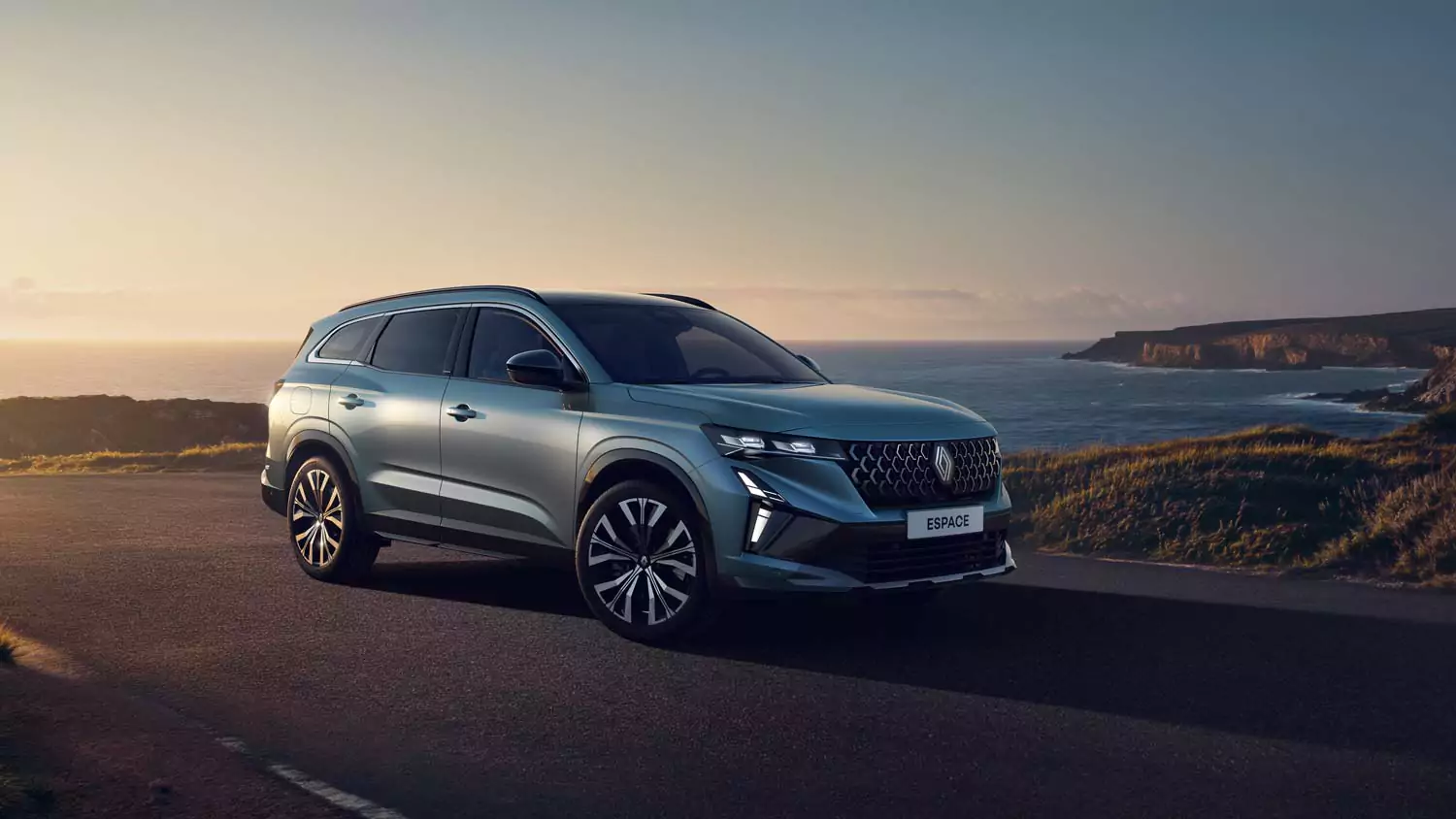 Ein silberner Geländewagen parkt bei Sonnenuntergang an einer Küstenstraße, mit Klippen, grasbewachsenen Hügeln und dem Meer im Hintergrund. Der Himmel ist klar und schafft eine ruhige und malerische Atmosphäre.