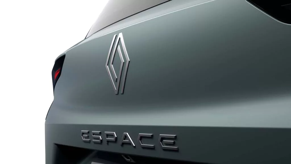 Nahaufnahme des Hecks eines Autos mit Metallic-Lackierung, auf dem das Renault-Logo und das Wort ESPACE in fetten silbernen Buchstaben zu sehen sind. Der Hintergrund ist weiß und ein Teil des Rücklichts ist sichtbar.