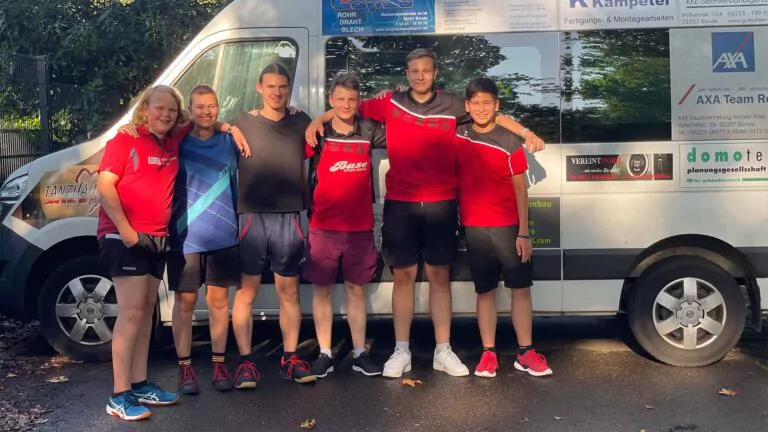 Sechs junge Leute stehen lächelnd und Arm in Arm vor einem weißen Lieferwagen mit verschiedenen Werbungen und Logos. Die meisten von ihnen tragen sportliche Kleidung, und das Sonnenlicht scheint von links auf sie.