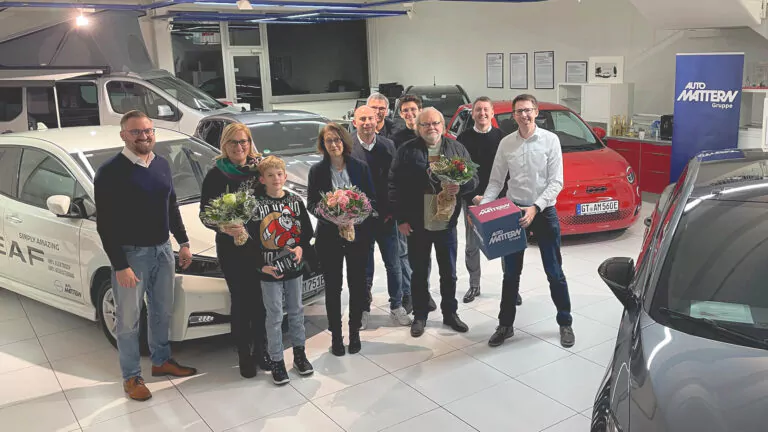 Eine Gruppe von Personen steht in einem Autohaus, einige halten Blumensträuße in der Hand. Im Hintergrund sind Autos zu sehen, und eine Person hält eine blaue Auto Matterw-Tasche. Alle lächeln und schauen in die Kamera.
