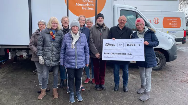 Eine Gruppe von Menschen in Winterkleidung steht draußen, lächelt und hält einen großen Scheck der Spende Tafel Mattern über 2.937 Euro für die Tafel Deutschland e.V. Im Hintergrund ist ein Lieferwagen mit Spendenaufschriften zu sehen.