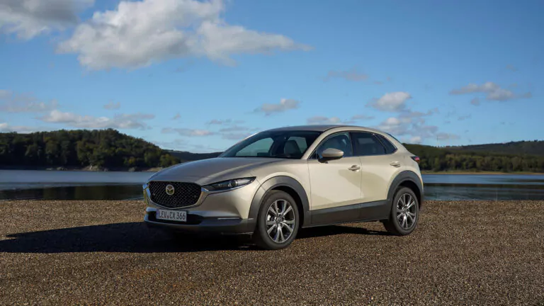 Ein silberner Mazda CX-30 Geländewagen parkt auf einer Kiesfläche in der Nähe eines ruhigen Sees, mit grünen Bäumen und Hügeln im Hintergrund unter einem blauen Himmel mit vereinzelten Wolken.