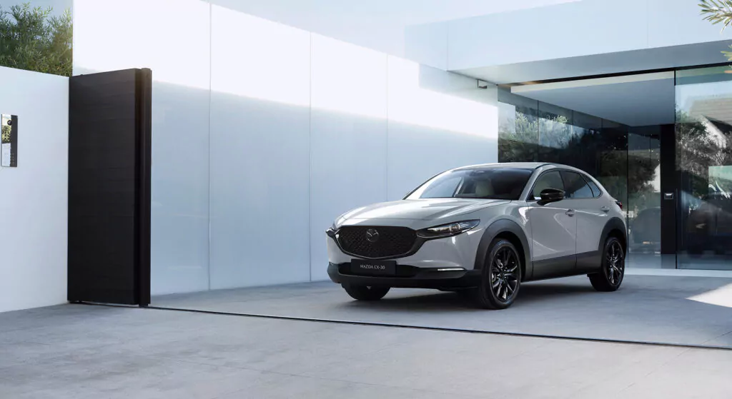Vor einem modernen, glasverglasten Haus mit einer Einfahrt und einem schwarzen Schiebetor steht ein silberner Mazda CX-30. Die Szene ist minimalistisch und gut ausgeleuchtet, wodurch das schnittige Design des Autos besonders zur Geltung kommt.