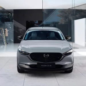 Ein silberner Mazda CX-30 steht vor einem modernen Gebäude mit Glasfassade; die Aufnahme zeigt ihn von vorne, wobei die Scheinwerfer, der Kühlergrill und das Emblem zu sehen sind.