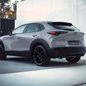 Ein grauer Mazda CX-30 SUV, der im Freien in der Nähe eines modernen Gebäudes geparkt ist, von hinten links gesehen. Das Fahrzeug hat schwarze Felgen und wird von sanftem Tageslicht beleuchtet.
