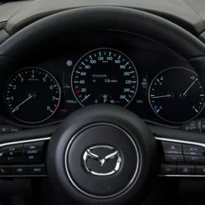 Nahaufnahme des Armaturenbretts eines Mazda-Fahrzeugs, auf dem das Lenkrad mit dem Mazda-Logo sowie drei beleuchtete Anzeigen zu sehen sind, die Geschwindigkeit, Kraftstoffstand, Temperatur und weitere Fahrdaten anzeigen.