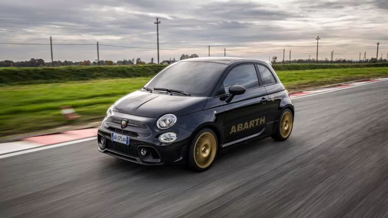 Ein schwarzer Abarth 695 Anniversario mit goldenen Rädern und Aufklebern rast über eine Rennstrecke, umrahmt von grünem Gras und einem bewölkten Himmel im Hintergrund.