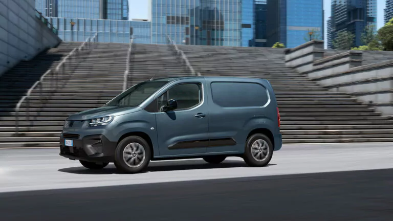 Ein grauer Lastwagen parkt an einem sonnigen Tag auf einer Straße vor modernen gläsernen Bürogebäuden und breiten Betontreppen.