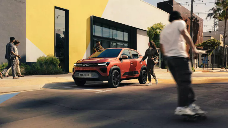 Ein roter Geländewagen der Kompaktklasse ist auf einer Straße in der Stadt geparkt, während drei Personen in der Nähe auf dem Bürgersteig stehen. Im Vordergrund fährt eine Person auf einem Skateboard vorbei, und im Hintergrund sind moderne Gebäude zu sehen.