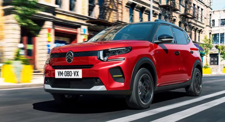 Ein roter Citroën-Geländewagen fährt zügig eine Stadtstraße entlang, die von modernen Gebäuden, gelben Blumenkübeln und verschwommenen Bewegungen gesäumt ist, die auf Geschwindigkeit und städtische Aktivität hindeuten.