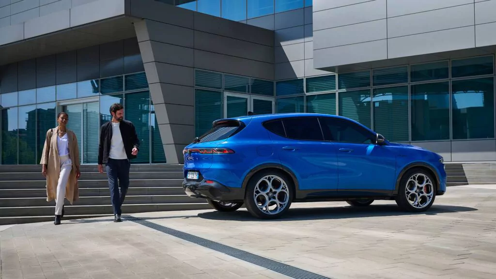 Ein Mann und eine Frau gehen eine Treppe in der Nähe eines modernen Glasgebäudes hinunter, während ein Alfa Romeo Tonale Speciale Geländewagen in der Nähe auf dem Pflaster geparkt ist.