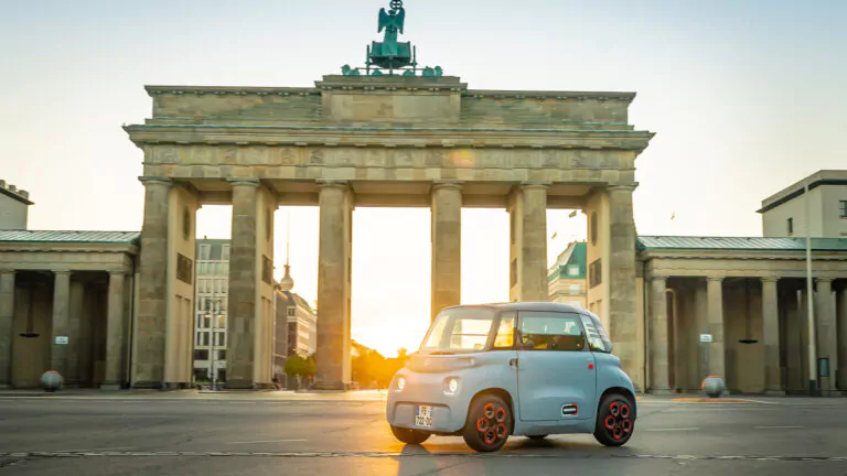 Ein kleines, hellblaues Elektroauto fährt bei Sonnenaufgang vor dem historischen Brandenburger Tor in Berlin, das von sanftem Sonnenlicht erhellt wird.