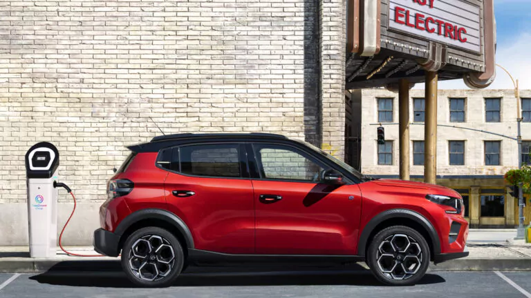 Ein roter Elektro-Geländewagen parkt neben einer Ladestation an einer Stadtstraße, angeschlossen und aufgeladen in der Nähe eines Backsteingebäudes mit einem teilweise sichtbaren Schild mit der Aufschrift ELECTRIC.