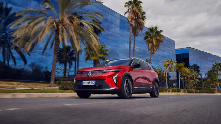 Ein roter Renault fährt auf einer von Palmen gesäumten Straße vorbei an modernen Glasgebäuden unter einem teilweise bewölkten Himmel.