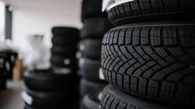 Nahaufnahme von gestapelten Autoreifen mit detaillierten Laufflächenmustern im Fokus, in einem Lagerhaus oder einer Werkstatt mit weiteren Reifenstapeln im unscharfen Hintergrund.