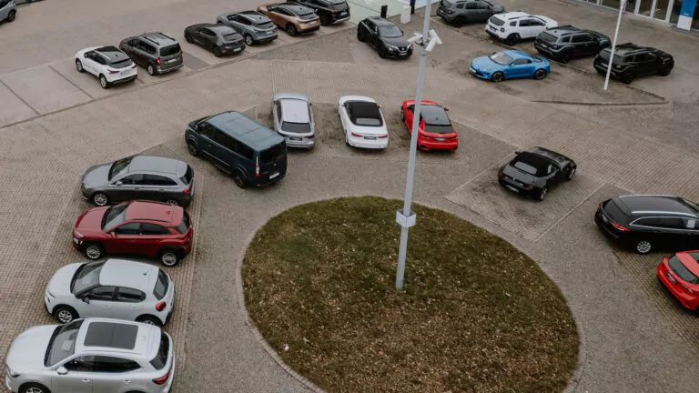 Luftaufnahme eines kreisförmigen Parkplatzes mit einer grasbewachsenen Insel in der Mitte, umgeben von verschiedenen geparkten Autos in unterschiedlichen Farben, darunter rot, weiß, silber, schwarz und blau.