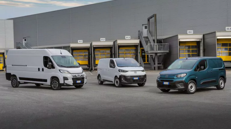 Drei weiße Lieferwagen parken vor einem modernen Lagerhaus mit mehreren Laderampen und gelben Türen. Die Szene wirkt industriell und organisiert, mit einem grauen Gebäude im Hintergrund.