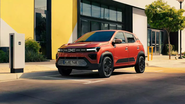 Ein roter Kompakt-SUV parkt neben einer Ladestation für Elektrofahrzeuge vor einem modernen Gebäude mit gelben und weißen Wänden und großen Fenstern. Ein Baum spendet in der Nähe Schatten.