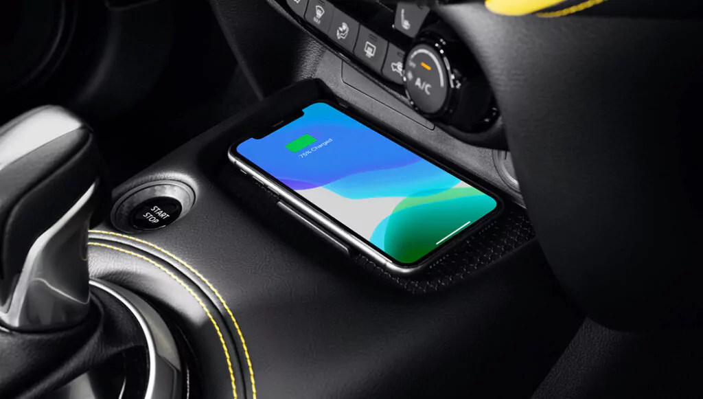 Ein Smartphone wird drahtlos auf einem Pad in der Mittelkonsole eines Autos aufgeladen, in der Nähe des Schalthebels, des Start-/Stopp-Knopfes und der Klimakontrollknöpfe. Auf dem Telefondisplay erscheint ein grünes Batteriesymbol, das den Ladevorgang anzeigt.