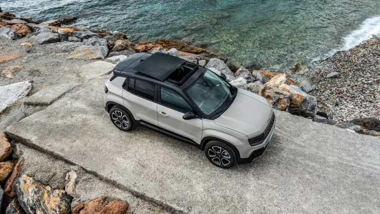 Ein beiger Geländewagen mit schwarzem Dach und geöffnetem Schiebedach ist auf einer Betonfläche in der Nähe der felsigen Ufer geparkt, mit Blick auf das klare blaugrüne Wasser.