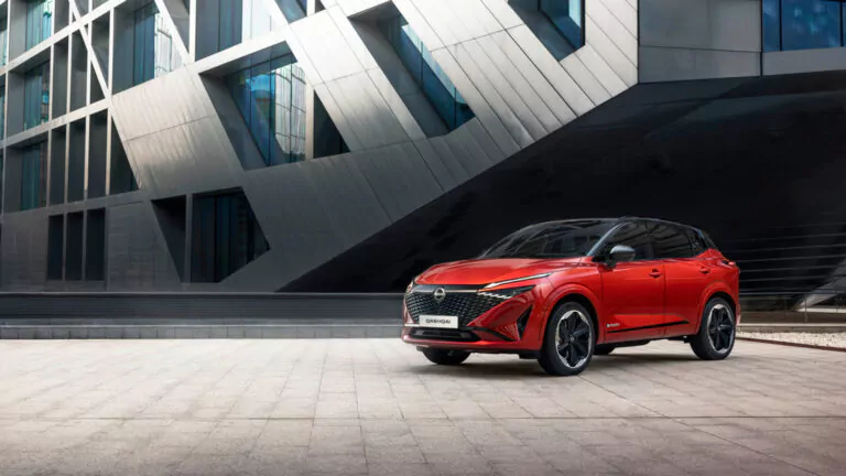 Ein roter Kompakt-SUV parkt auf einem modernen, minimalistischen Steinplatz vor einem zeitgenössischen Gebäude mit kantigen Fenstern und geometrischen Metallplatten.