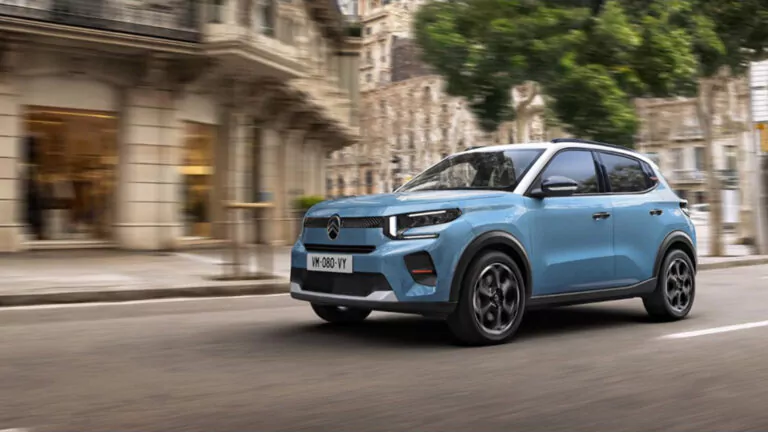 Ein hellblauer Citroën-Geländewagen fährt auf einer Stadtstraße mit unscharfen Gebäuden und grünen Bäumen im Hintergrund, die Bewegung suggerieren.