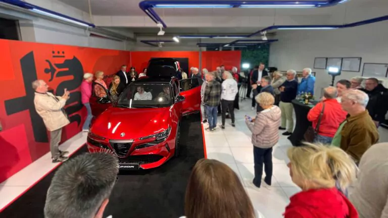 Während der exklusiven Vorpremiere von Alfa Romeo Junior in einem hell erleuchteten Ausstellungsraum versammelt sich eine Menschenmenge um einen leuchtend roten Alfa Romeo Geländewagen. Einige machen Fotos, andere bewundern das Fahrzeug aus der Nähe.