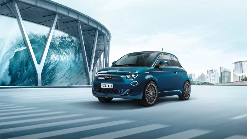 Ein blauer Fiat 500 Elektro fährt durch eine moderne Straße mit futuristischen Gebäuden, vorbei an einem großen Wellenkunstwerk auf einer geschwungenen Struktur - perfekt für alle, die Wallbox-Leasingoptionen in Betracht ziehen.